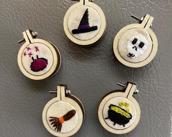 Puede incluir: Una colección de cinco pequeños aros de bordado redondos de madera. Cada aro presenta un diseño bordado diferente con temática de Halloween, que incluye un sombrero de bruja, una calavera, un caldero, una escoba y un racimo de bayas.