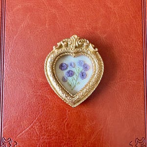 May include: A heart-shaped, gold-colored frame with an embroidered floral design. The frame has ornate detailing and surrounds a cream-colored fabric with purple flowers. The background is a textured, reddish-brown surface.