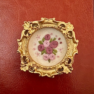 May include: A small, gold-colored ornate frame with an embroidered floral design. The embroidery features a bouquet of pink and purple roses with green leaves on a cream-colored background. The frame is set against a textured, reddish-brown surface.