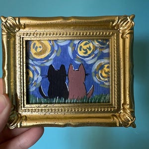 May include: A small framed painting featuring two cat silhouettes, one black and one brown, gazing at a starry night sky. The artwork is set in a gold ornate frame. The background is a blue sky with yellow swirls.