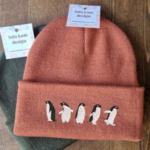 May include: Two knit beanies, one rust-colored and one olive green, are stacked on a wooden surface. The rust-colored beanie features an embroidered design of five penguins. Each beanie has a tag that reads "lulu kate design".