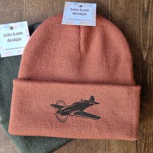 May include: Two beanies, one rust-colored and one olive green, are stacked on a wooden surface. The rust-colored beanie features a black airplane graphic. Each beanie has a tag that reads "lulu kate design".