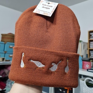 May include: A rust-coloured knit beanie featuring an embroidered design of four white geese in various poses on the folded cuff. A white rectangular tag attached to the top of the beanie displays the text lulu kate design in black font.
