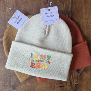 May include: Three beanies in shades of tan, beige, and rust are stacked on a wooden surface. The frontmost beanie is beige with the embroidered text "IN MY SOURDOUGH ERA" in colorful thread. Each beanie has a tag that reads "lulu kate design".