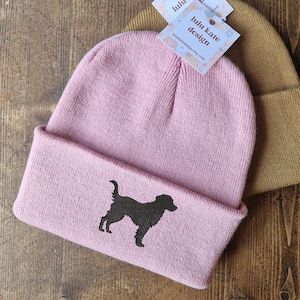以下が含まれることがあります： 木製の表面に、ピンクとタンの 2 つのニット帽が表示されています。 ピンクの帽子には、犬の黒いシルエットがあります。 添付のタグには「lulu kate design」と書かれています。
