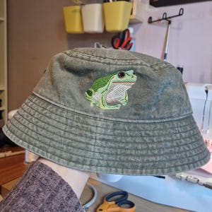 May include: A green bucket hat with a detailed embroidered frog design. The hat is a faded green color and has a classic brim. The frog is green and white, adding a pop of color to the hat.