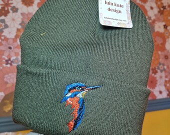 May include: An olive green knit beanie featuring an embroidered kingfisher bird on the front cuff. The bird has blue, orange, and white thread details. A white rectangular tag attached to the top reads lulu kate design and lulukatedesign.etsy.com.