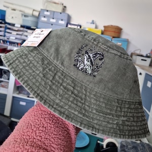 May include: Olive green bucket hat with an embroidered mushroom design. The hat has a structured brim and a tag that reads "Julia Kate Design". The hat is made of a soft, textured fabric.