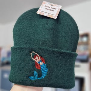May include: A dark green knit beanie featuring an embroidered mermaid with red hair and a blue tail. A white tag at the top displays the text lulu kate design and lulukatedesign.etsy.com. The beanie has a folded cuff and a textured knit fabric.