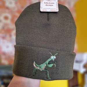May include: An olive green knit beanie featuring an embroidered green praying mantis on the front cuff. A white rectangular tag at the top displays the text lulu kate design and lulukatedesign.etsy.com in a brown serif font with a floral border.