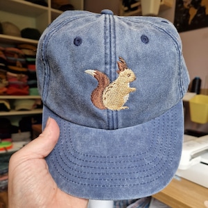 May include: A blue baseball cap featuring an embroidered squirrel. The squirrel is rendered in brown and tan hues, with a bushy tail. The cap has a curved peak and a fabric strap at the back for adjustment.