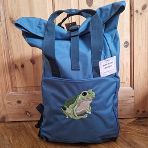 Könnte beinhalten: Türkisfarbener Rucksack mit einer grünen Froschstickerei auf der Vordertasche. Er hat einen oberen Griff und verstellbare Träger. Das Etikett lautet "lulu kate design".