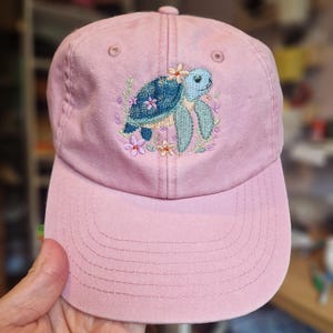 May include: A pale pink baseball cap with an embroidered sea turtle design. The turtle has a blue shell and light blue limbs, surrounded by small flowers. The cap has a curved peak and a fabric strap.