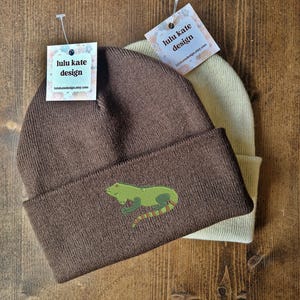 May include: Two knitted beanies, one brown and one cream, are shown. The brown beanie has a green lizard embroidery. Both beanies have tags that read "lulu kate design" and the website address.