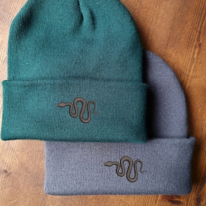 May include: Two knit beanies, one green and one grey, with a black snake embroidered on the front of each.