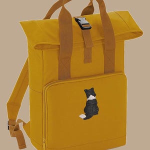 May include: Mustard yellow backpack with a roll-top closure and a brown handle. The front pocket features an embroidered black and white dog. The backpack has brown straps and a black buckle.