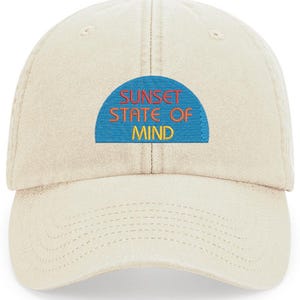 May include: A beige baseball cap featuring the embroidered phrase "SUNSET STATE OF MIND" in orange and yellow lettering on a blue patch. The cap has a curved peak and a structured crown.
