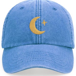 May include: A light blue baseball cap with a curved peak. The cap has an embroidered gold crescent moon and star design on the front. The cap is made of a soft, washed fabric and has an adjustable strap.