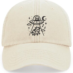 May include: A beige baseball cap featuring a black graphic of an alien in a spaceship abducting a cow. The design includes stars, a crescent moon, and musical notes. The cap has a curved peak and an adjustable strap.