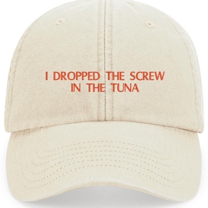 May include: A light beige baseball cap with the embroidered text "I DROPPED THE SCREW IN THE TUNA" in red. The cap has a curved brim and a fabric strap closure.