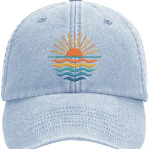 May include: A light blue denim baseball cap featuring an embroidered graphic of a sun rising over ocean waves. The sun is orange with yellow rays, and the waves are depicted in horizontal layers of orange, yellow, teal, and dark blue thread.
