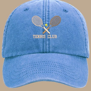 May include: A blue denim baseball cap with a white embroidered design of two crossed tennis rackets and a tennis ball. The text "Tennis Club" is embroidered in white below the rackets.