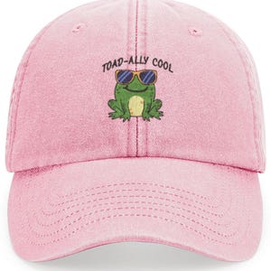 May include: A light pink baseball cap with a curved brim. The cap features an embroidered graphic of a green frog wearing sunglasses, with the text "TOAD-ALLY COOL" above it. The cap is made of a soft fabric.