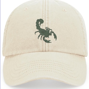 May include: A cream-coloured baseball cap with a dark green scorpion embroidered on the front. The cap has a curved peak, a top button, and stitched details. The adjustable strap is not visible.