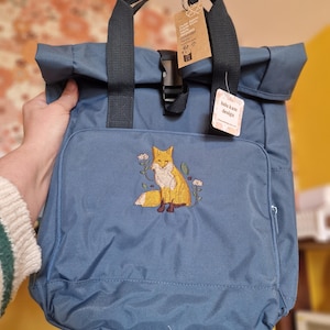 May include: A slate blue roll-top backpack featuring an embroidered orange fox surrounded by small flowers on the front pocket. The bag has black nylon straps, a plastic buckle closure, and two paper tags attached, one reading lulu kate design.
