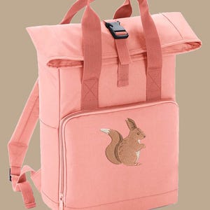 May include: A coral-coloured backpack with a roll-top closure, featuring a brown embroidered squirrel on the front pocket. The backpack has adjustable shoulder straps, a top handle, and a black buckle closure. The bag is made of a durable fabric.
