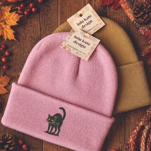 May include: A light pink knit beanie featuring an embroidered black cat with orange eyes on the cuff, accompanied by a mustard yellow knit beanie. Both beanies have a square paper tag attached that reads lulu kate design in black text.