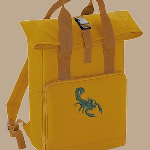May include: Mustard yellow backpack with a roll-top design and brown straps. A teal scorpion is embroidered on the front pocket. Features a top handle and adjustable shoulder straps. Includes a zippered front pocket and a black buckle.