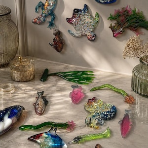 Può includere: Una collezione di colorati oggetti d'arte in resina di varie forme, tra cui pesci, creature marine e forme astratte. I pezzi sono esposti su una superficie di marmo, alcuni montati su una parete. I colori includono blu, verde, rosa e oro.
