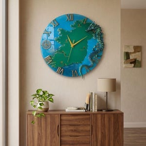 May include: A round wall clock featuring a map of Ireland in green, surrounded by a blue ocean. Gold Roman numerals mark the hours. A green dragon decorates the right side. The clock is mounted on a beige wall.