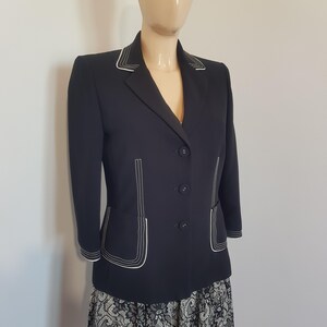 Può includere: Un blazer blu navy scuro con tre bottoni frontali, maniche a tre quarti e cuciture bianche a contrasto lungo i revers, i polsini e le due tasche anteriori. Il blazer è indossato sopra una gonna con motivo in bianco e nero.