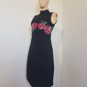 Può includere: Un abito nero senza maniche con collo alto e un'applicazione floreale decorativa sul petto. Il motivo floreale è composto da petali rosa e rossi con foglie verdi, arricchito da paillettes per una finitura leggermente riflettente.