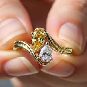 May include: A gold ring with two pear-shaped gemstones. One is yellow, the other clear. The ring has a flowing, wave-like design. The ring is held between two hands.
