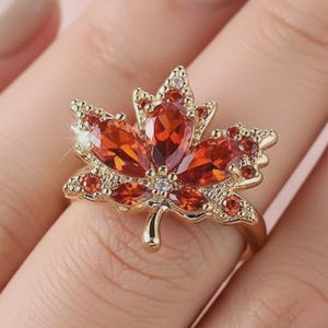 Könnte beinhalten: Ein goldfarbener Ring mit einem Ahornblatt-Design. Das Blatt besteht aus orangefarbenen und roten Edelsteinen, mit kleineren klaren Steinen an den Rändern. Der Ring befindet sich vor einem zartrosa Hintergrund.