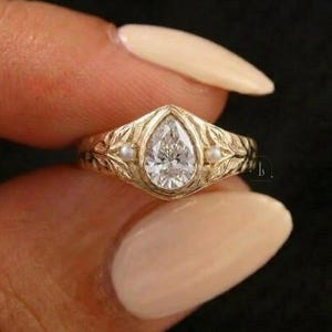 May include: A gold ring with a pear-cut diamond centre stone. The ring has a detailed floral design on the band and small pearl accents either side of the diamond. The ring is held between fingers against a dark background.