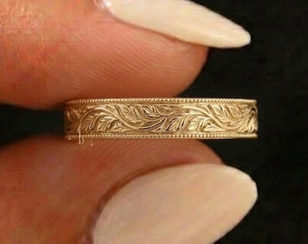 May include: A gold band ring featuring an intricate, continuous engraved leaf pattern along the exterior surface. The metal has a warm, polished finish, and the design is bordered by fine, beaded detailing along both the top and bottom edges.