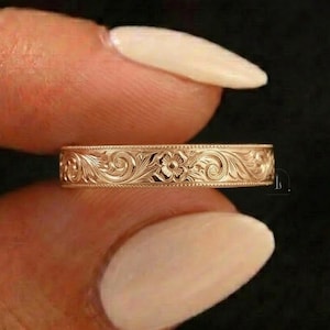 May include: A rose gold band ring featuring an intricate engraved floral pattern with swirling scrollwork details. The metal surface has a polished finish, and the edges of the band are lined with a fine beaded texture throughout the circumference.