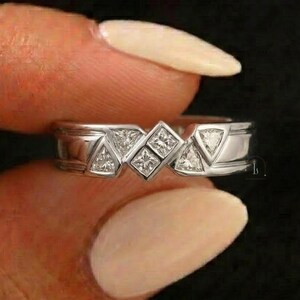 May include: A silver ring with a geometric design featuring square and triangular-shaped gemstones. The ring has a polished finish and is held against a dark background.