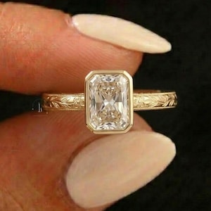 Peut inclure: Une bague en or avec une pierre précieuse rectangulaire et transparente. La bague a un sertissage à lunette et un motif floral détaillé sur le bracelet. La bague est tenue sur un fond sombre.
