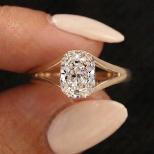 Puede incluir: Un anillo de oro con una piedra preciosa central de corte cojín con superficie facetada. La banda tiene un diseño dividido que se estrecha al conectar con el engaste que sujeta la piedra. El metal tiene un acabado pulido y tono dorado.