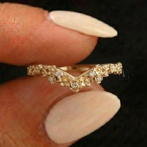 Può includere: Un anello color oro con un design floreale. L'anello presenta piccoli fiori e gemme scintillanti. L'anello è tenuto tra le dita su uno sfondo scuro. Il design dell'anello è a forma di V.