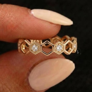 Pu&ograve; includere: Un anello color oro con un design a nido d'ape. L'anello presenta forme esagonali, alcune delle quali contengono piccole pietre preziose rotonde e trasparenti. L'anello &egrave; tenuto tra le dita su uno sfondo scuro.