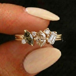 May include: A gold ring featuring a cluster of gemstones. The ring has a marquis-cut olive-green gemstone, a pear-shaped champagne-colored stone, and an emerald-cut white stone, along with smaller round diamonds. The band has a delicate, textured design.