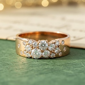 May include: A gold ring with a cluster of round, clear diamonds. The band is wide and features smaller diamonds and decorative accents. The ring is set against a green surface with a blurred background.
