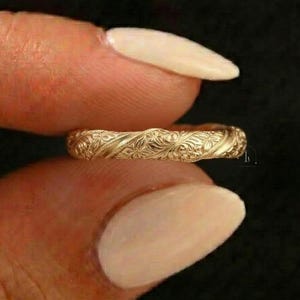 Puede incluir: Un anillo dorado con un intrincado diseño floral. El anillo se sostiene entre dos dedos sobre un fondo oscuro. El diseño del anillo presenta grabados detallados de flores y hojas, creando una superficie texturizada.