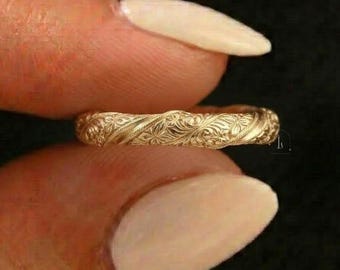 Könnte beinhalten: Ein goldfarbener Ring mit einem aufwendigen Blumenmuster. Der Ring wird zwischen zwei Fingern vor einem dunklen Hintergrund gehalten. Das Design des Rings zeigt detaillierte Schnitzereien von Blumen und Blättern, wodurch eine strukturierte Oberfläche entsteht.